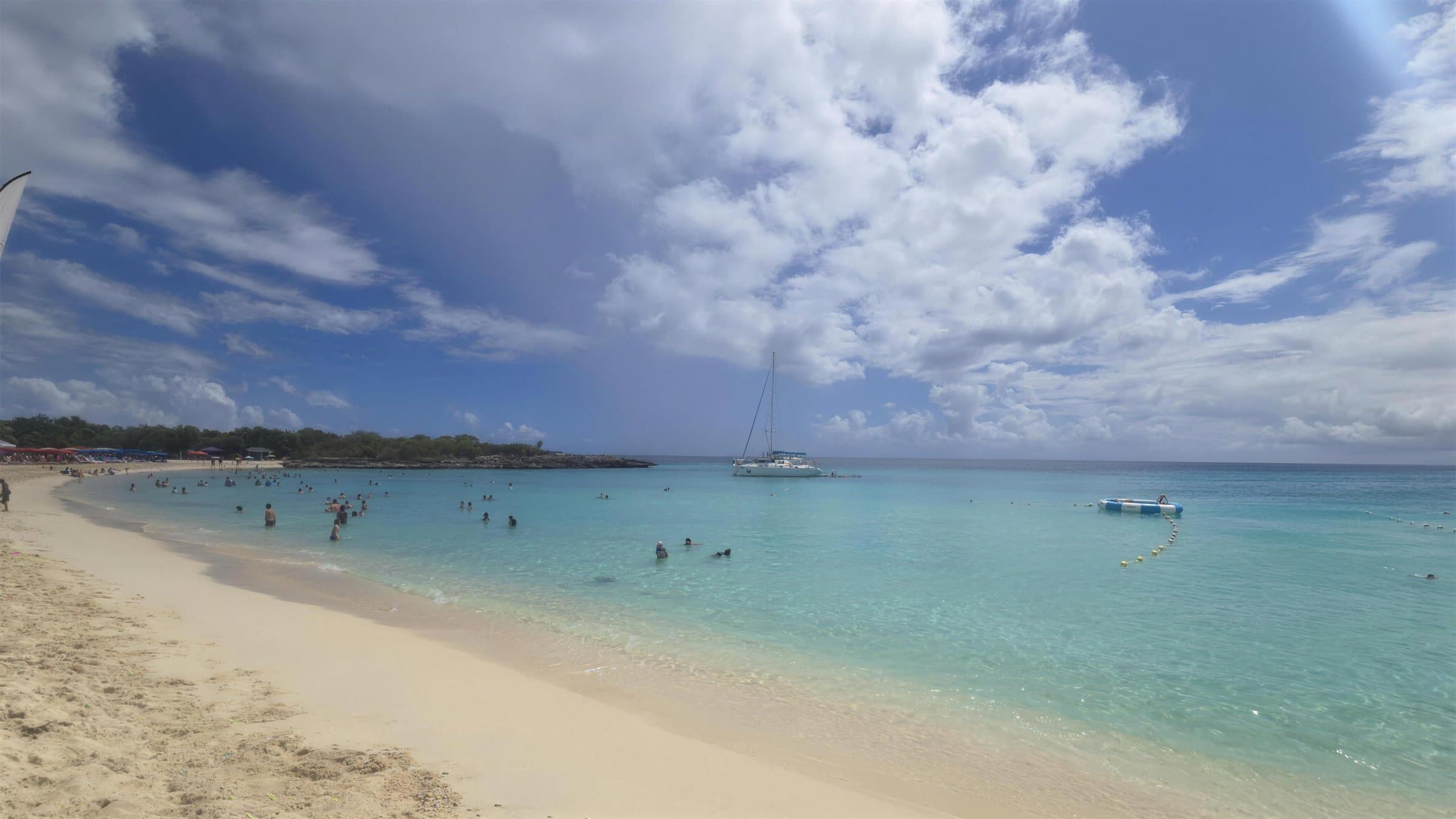 Eau turquoise à Mullet Bay, avec des baigneurs, un catamaran au mouillage et la pointe rocheuse au loin